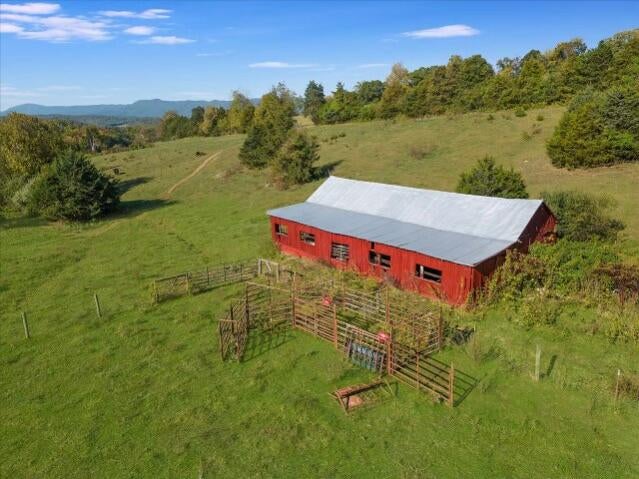 Aerial view of a rustic red barn surrounded by green pastures and trees, with a fenced area in the foreground, set against a backdrop of rolling hills and a clear blue sky.