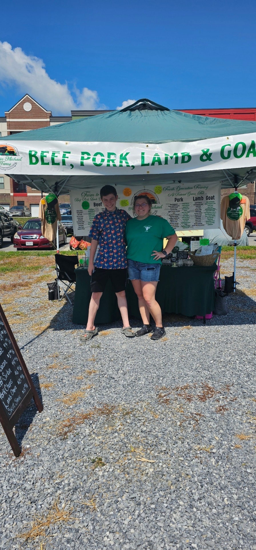 Farmers at a market stand showcasing beef, pork, lamb, and goat products, with a sign highlighting their offerings. The setting features a sunny day and a tent displaying fresh farm goods.