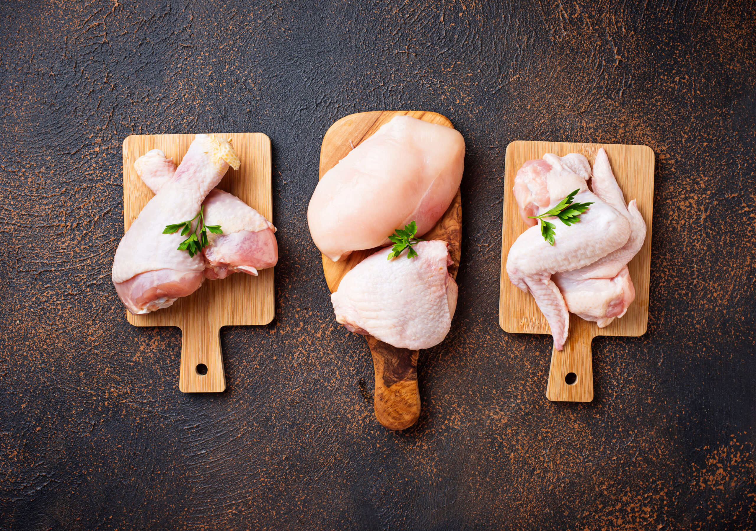 Raw chicken meat fillet, thigh, wings and legs Raw chicken pieces displayed on wooden cutting boards, including drumsticks, chicken breasts, and wings, garnished with fresh parsley, set against a dark textured background.