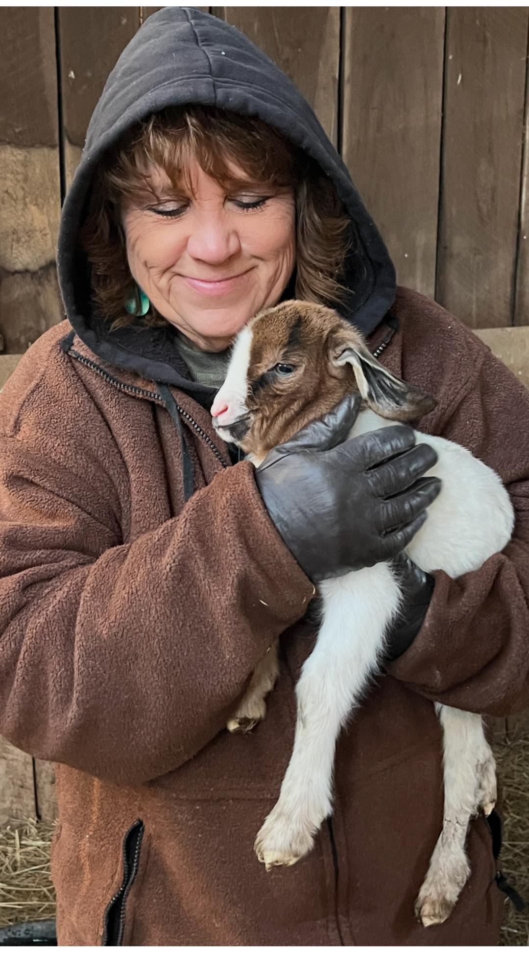 Woman in a brown jacket and gloves gently holds a baby goat, smiling affectionately. The background features wooden barn walls, indicating a farm setting.