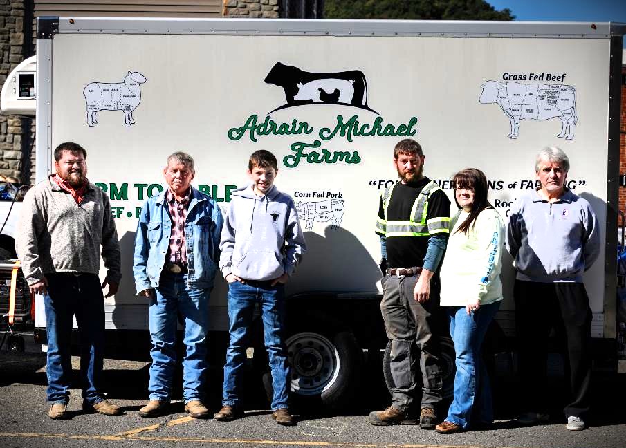 A group of six farmers stands in front of the Adrain Michael Farms trailer, showcasing their commitment to sustainable agriculture. The trailer features information about grass-fed and corn-fed beef, emphasizing farm-to-table practices. The farmers are dressed in casual attire, reflecting a hands-on approach to farming.