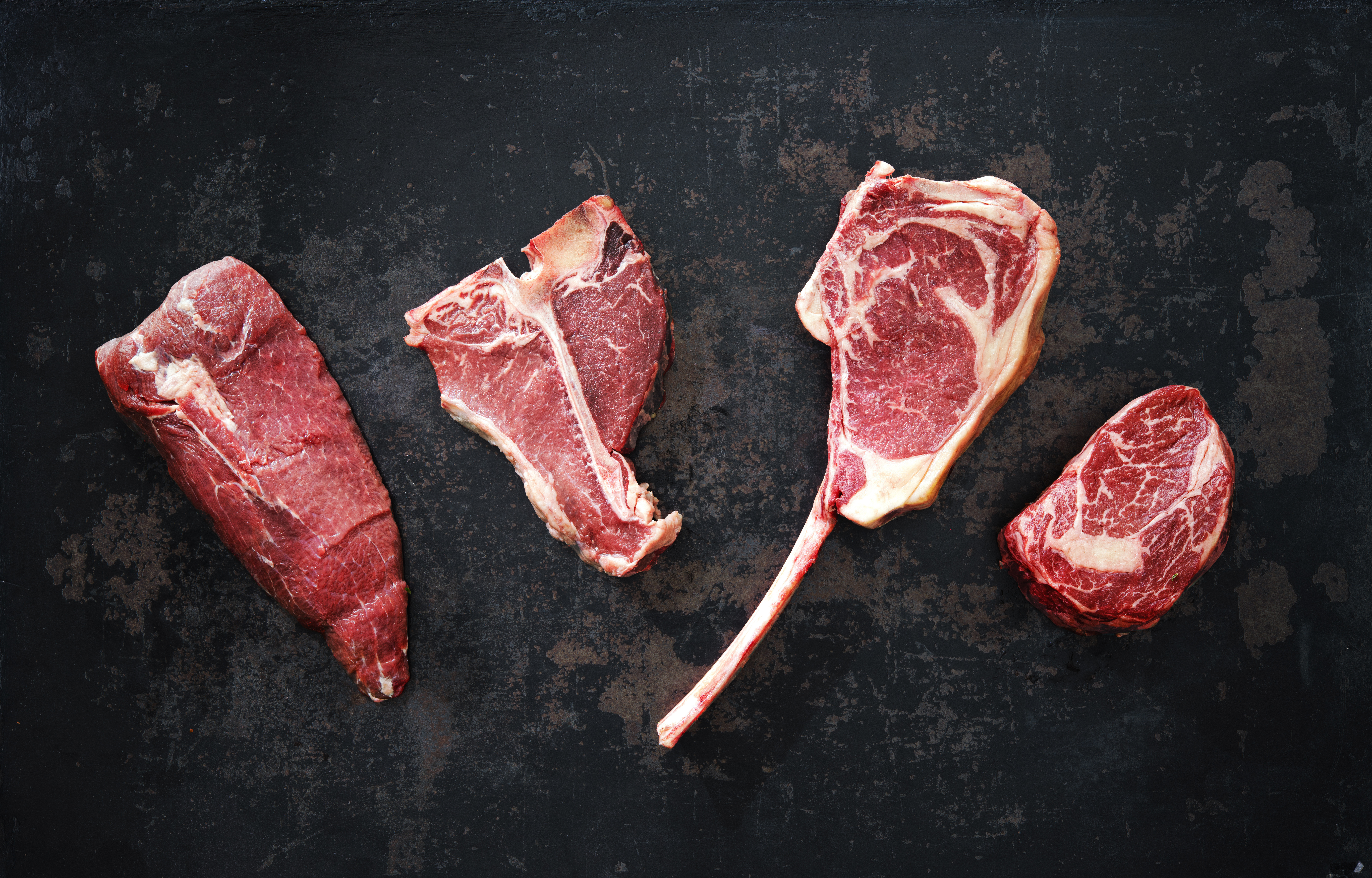 Variety of raw beef meat for grilling Various cuts of raw beef arranged on a dark surface, showcasing ribeye, T-bone, and filet mignon steaks, emphasizing marbling and freshness for meat enthusiasts.