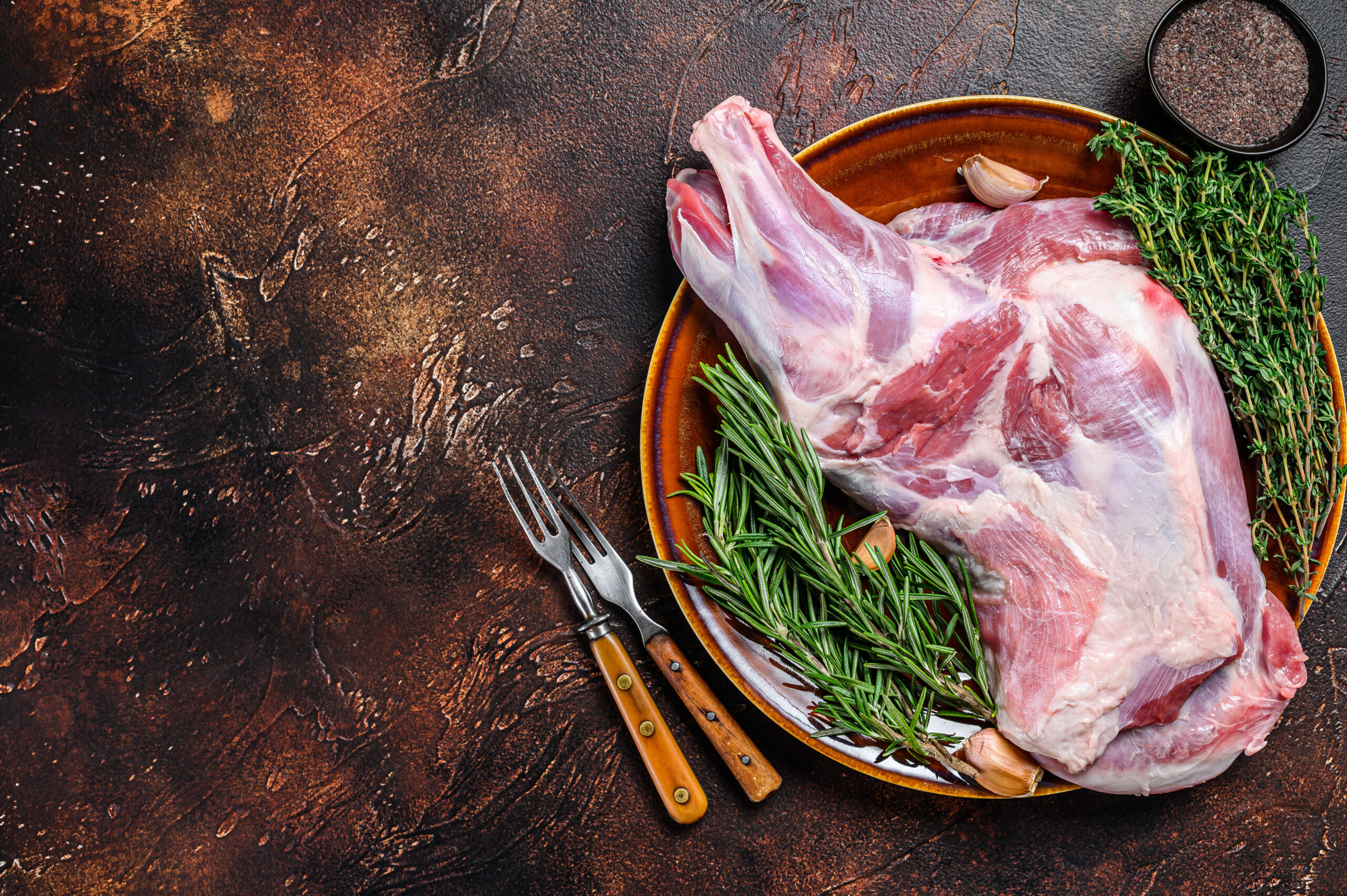 Whole fresh Raw goat shoulder meat on a plate. Dark wooden background. Top view. Copy space Raw lamb leg on a rustic plate, garnished with fresh rosemary, thyme, and garlic cloves, accompanied by two vintage forks, set against a textured dark background.