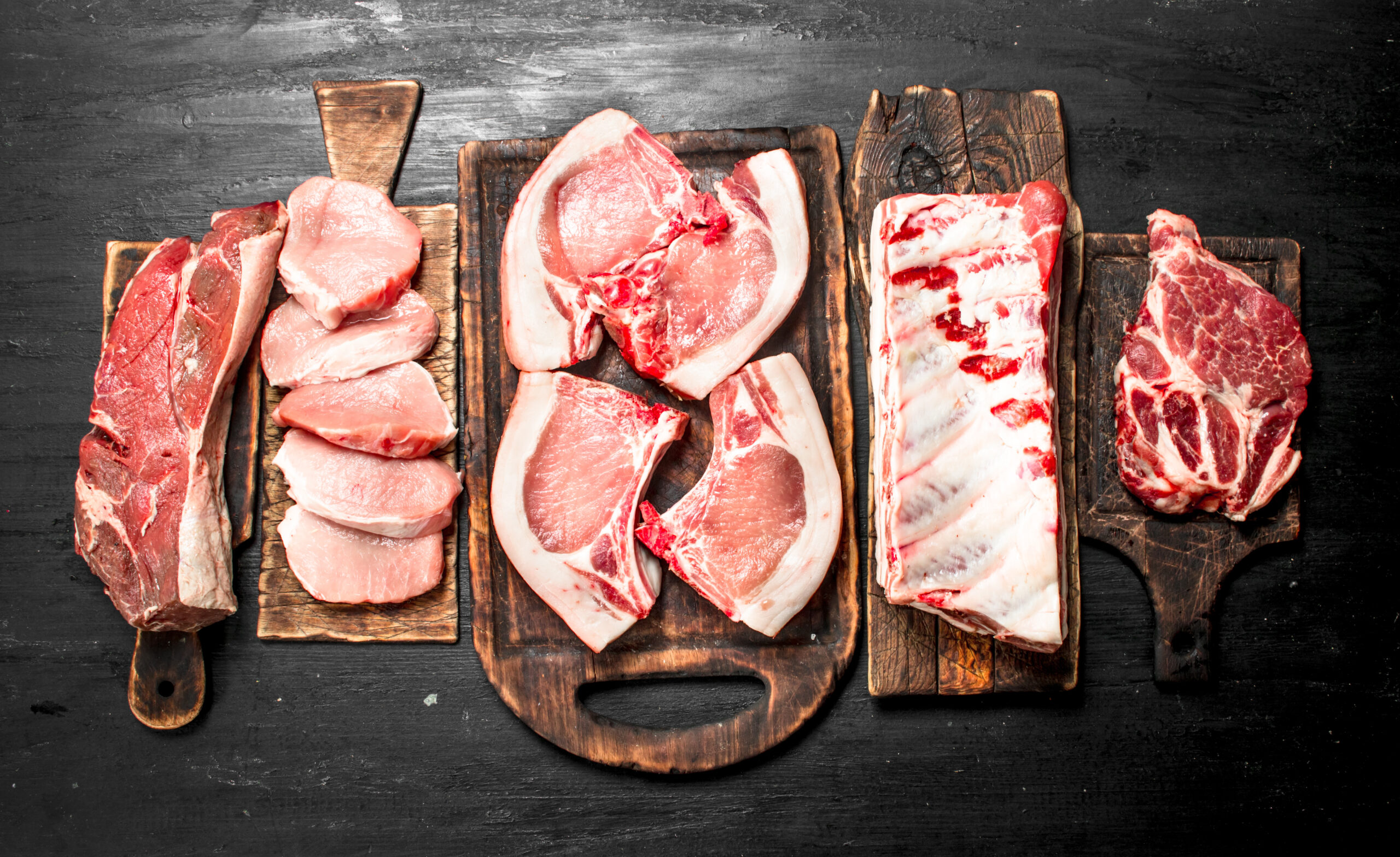 Different types of raw pork meat and beef. Assorted cuts of raw meat displayed on wooden cutting boards, including beef steaks, pork chops, and ribs, set against a dark background. Ideal for culinary, butchery, or food preparation contexts.